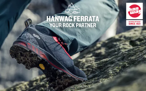 Close-up van een persoon die Hanwag Ferrata-wandelschoenen draagt en op een rotsachtige ondergrond loopt, waarbij het merklogo en de tekst ‘Hanwag Ferrata – Your Rock Partner’ zichtbaar zijn.